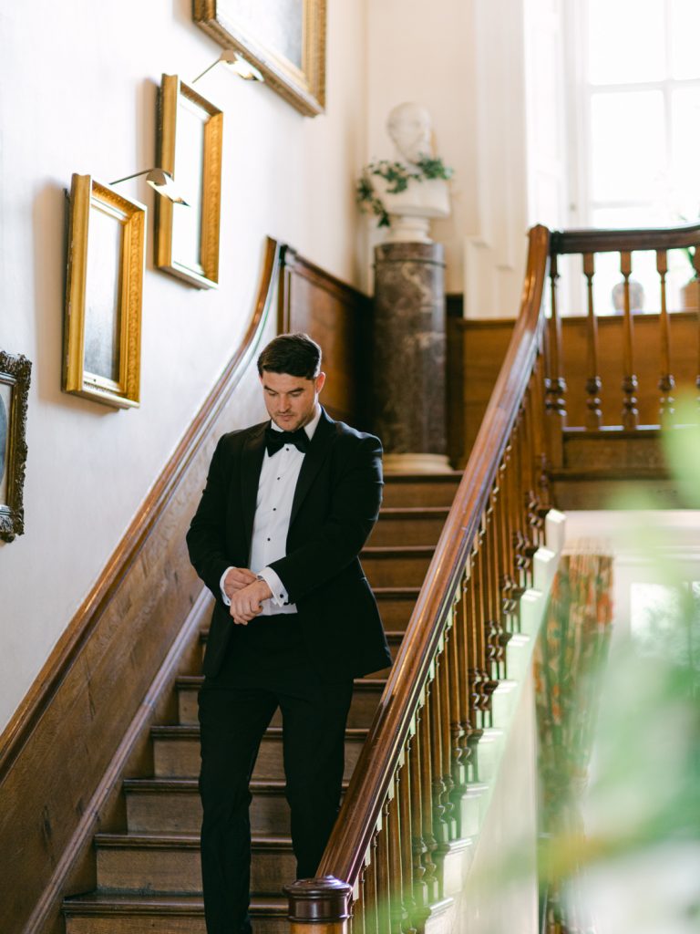 Groom Ryan dressed in black tie, fixing his cuff link whilst walking down the venue stairs to the ceremony