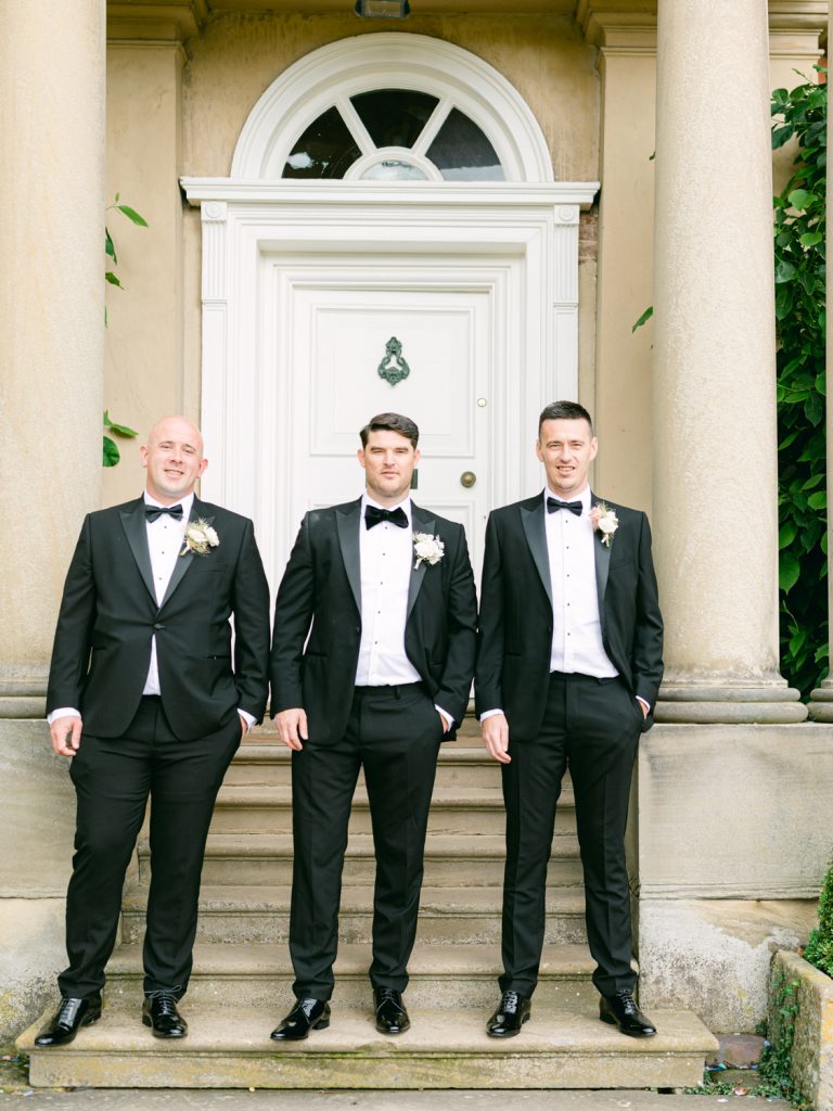 Groom Ryan and his best men standing together outside the front of the wedding venue, dressed in black tie.