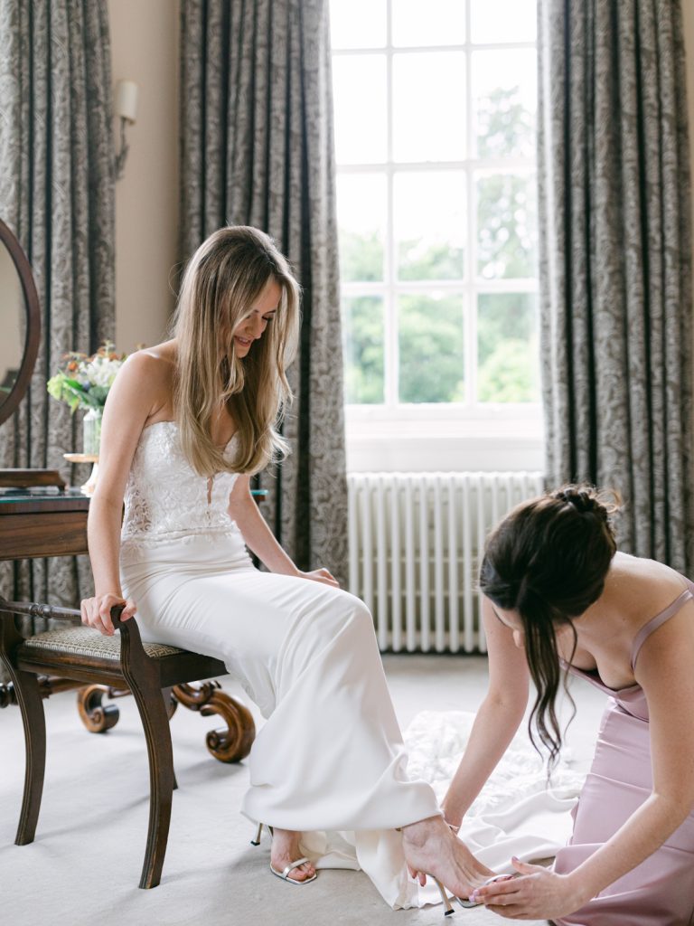 Bride Erin's bridesmaid helping her put her bridal shoes on during wedding prep.