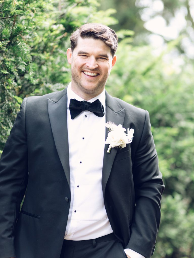 Groom Ryan on his own in the garden, wearing black tie and smiling broadly.