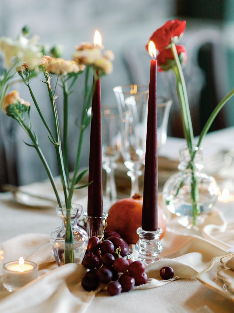 Detail shot of the tablescape including fruit for decoration, textured linens and dark red, lit tapered candles.