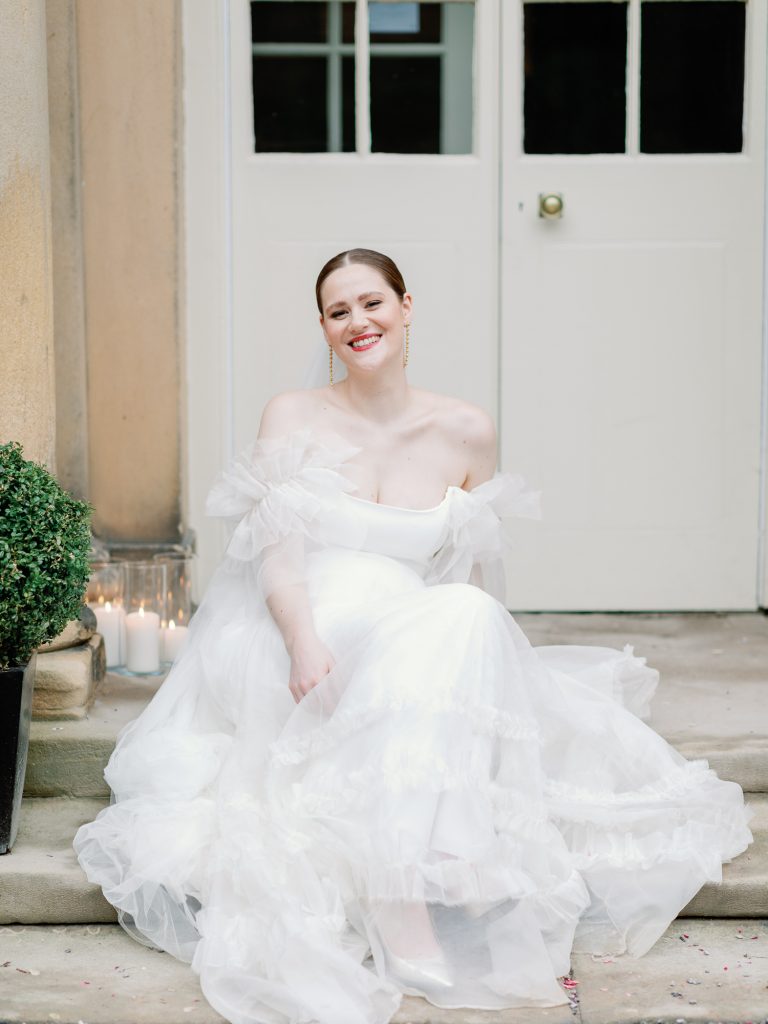 Bride Else sitting on the front steps, smiling at the camera with her tulle Halfpenny London gown gathered around her.