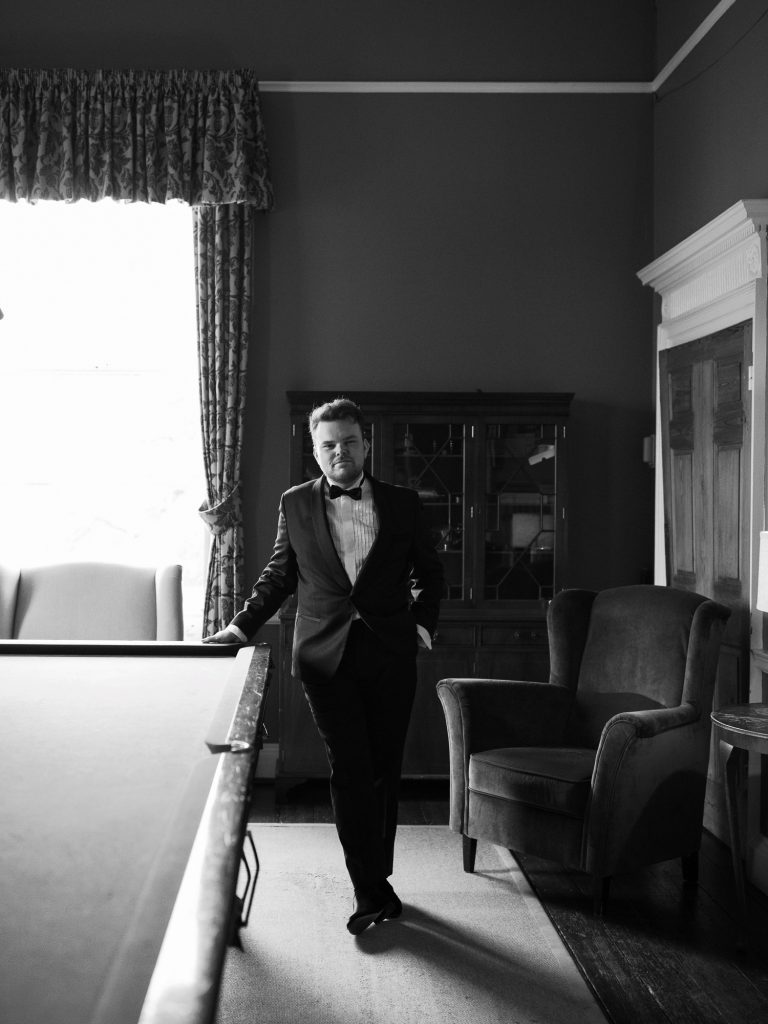 Groom Rory dressed in black tie leaning casually on a snooker table at Middleton Lodge.